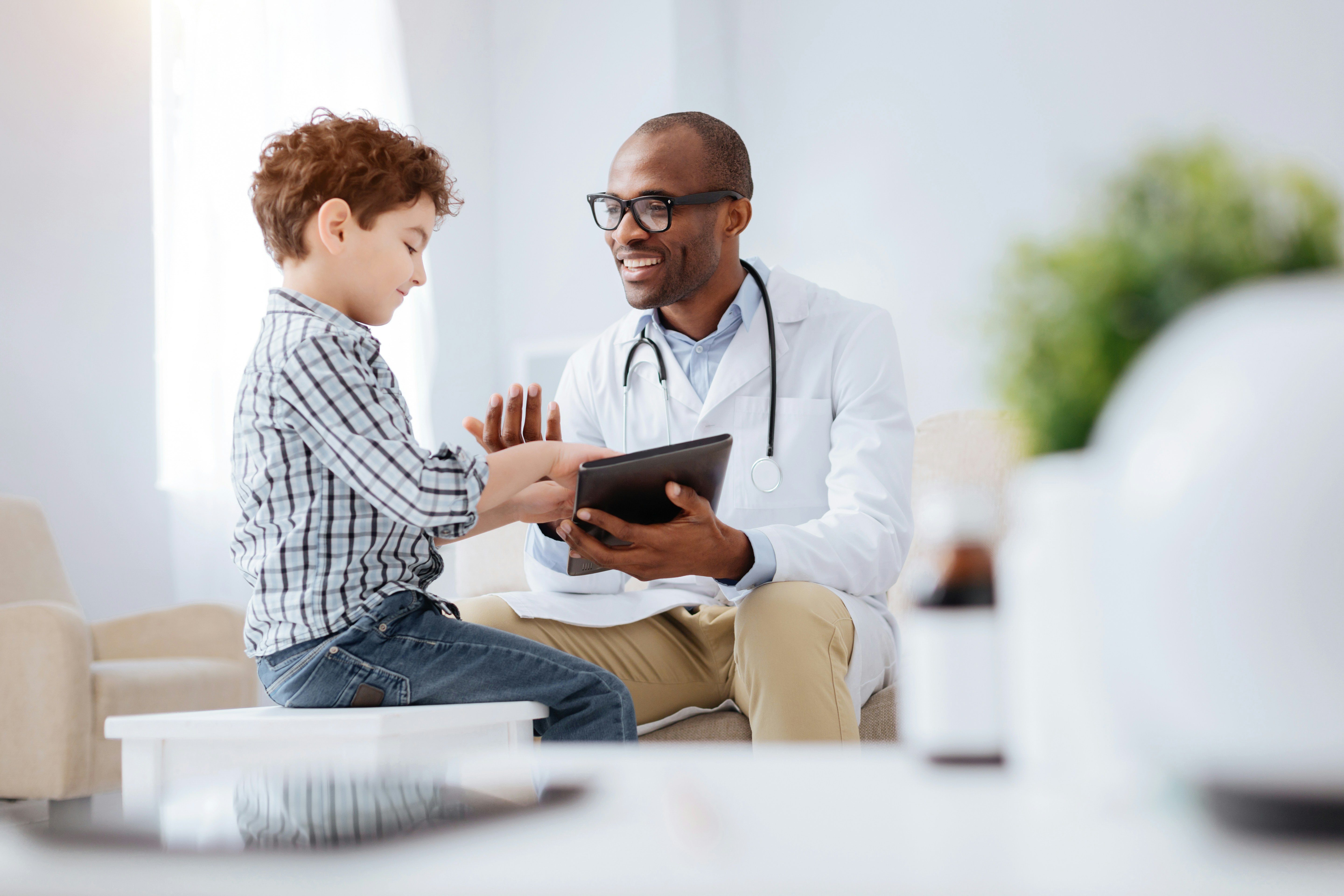 A doctor smiles with a young patient who taps the doctor's tablet.