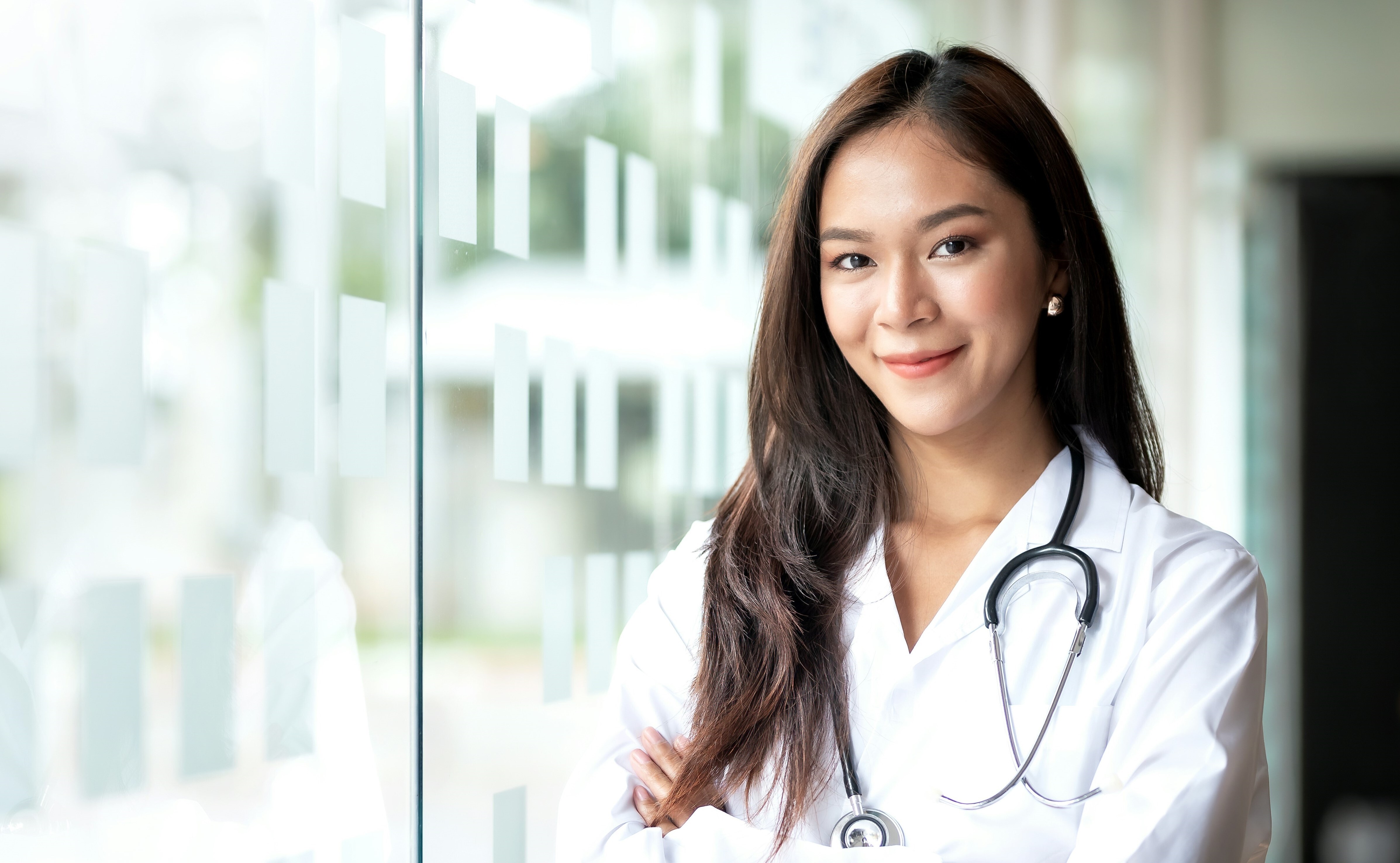 A young, female doctor stands near a window smiling at the camera with crossed arms.