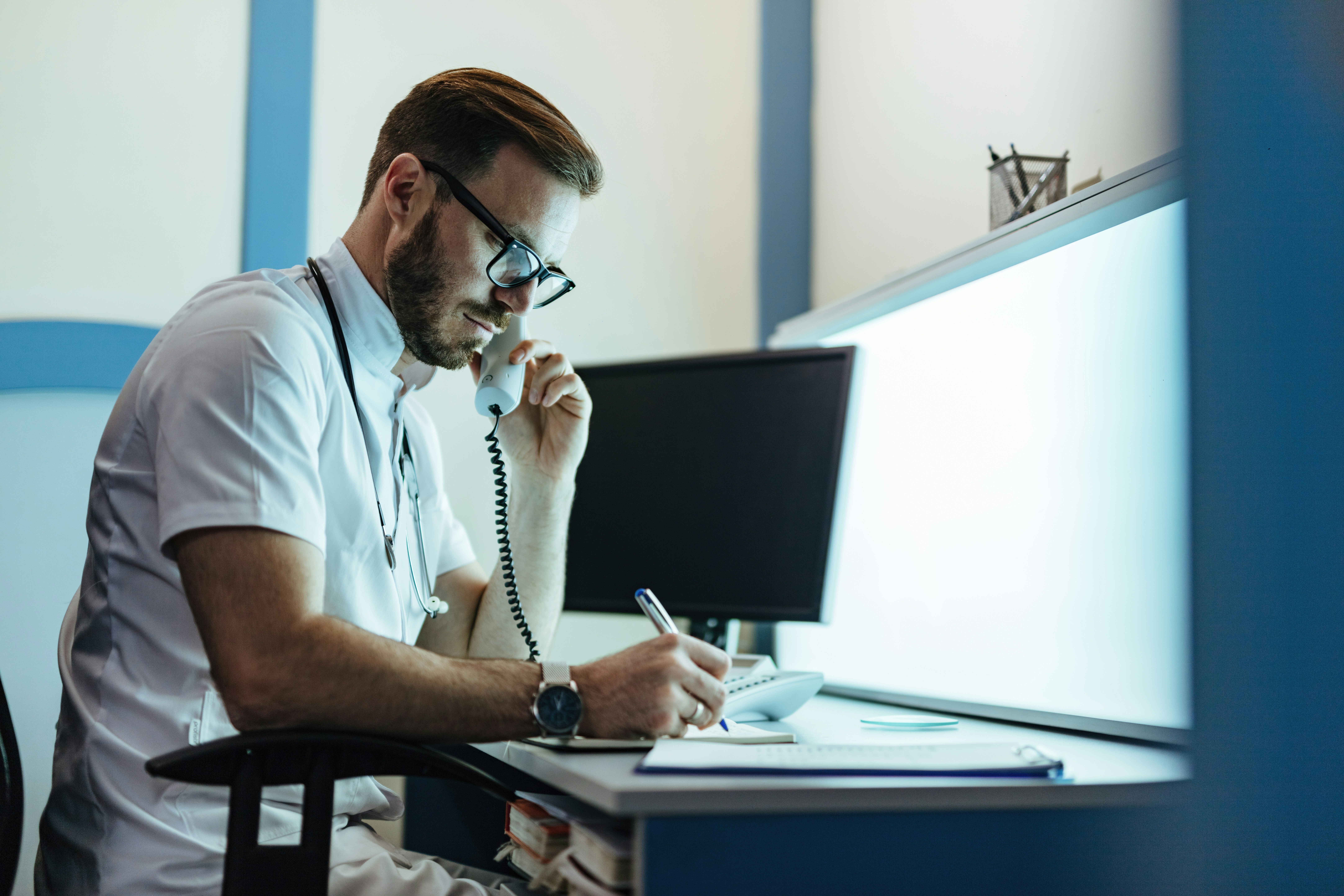 A doctor writes notes at a desk while speaking on the phone.